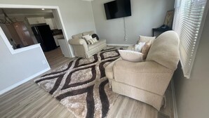 A living room with a beige sofa, a patterned rug, a flat-screen TV on the wall, and a window with blinds. - Charming 3-bedroom bungalow in pleasant Indianapolis with WiFi, AC, (Indianapolis)