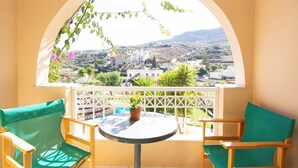 A balcony with a round table and two chairs, a potted plant, and a view of a town and hills. - Aquarius Apartments by Checkin (Agia Pelagia)