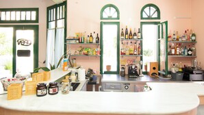 A kitchen with a coffee machine, shelves with bottles, and a window with curtains. - Aquarius Apartments by Checkin (Agia Pelagia)