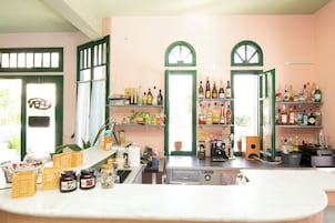 Una cocina con una cafetera, estantes con botellas y un ventanal con cortinas.