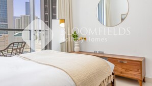 Un dormitorio con una cama grande, un aparador de madera, un espejo redondo y vistas de la ciudad a través de grandes ventanales.