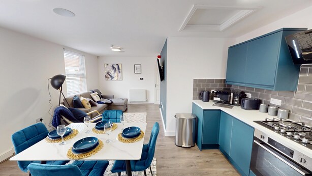 A compact kitchen with blue cabinets and a white countertop, a dining area with blue chairs and a table set for four, and a living area with a sofa and a coffee table. - For Workers | 10 Mins to City | 5 TVs and Parking (Liverpool)