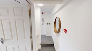 A hallway with white walls, a wooden-framed mirror, a red fire alarm, and a patterned carpet. - For Workers | 10 Mins to City | 5 TVs and Parking (Liverpool)