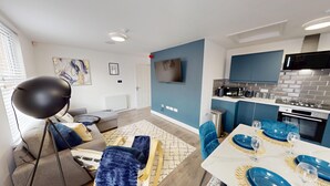 A modern living room with a sofa, a dining table set with blue plates and glasses, a kitchen with blue cabinets and a TV mounted on the wall. - For Workers | 10 Mins to City | 5 TVs and Parking (Liverpool)