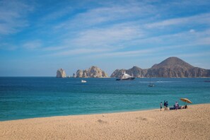 A beach with clear turquoise water, a yacht, and rocky formations in the distance.