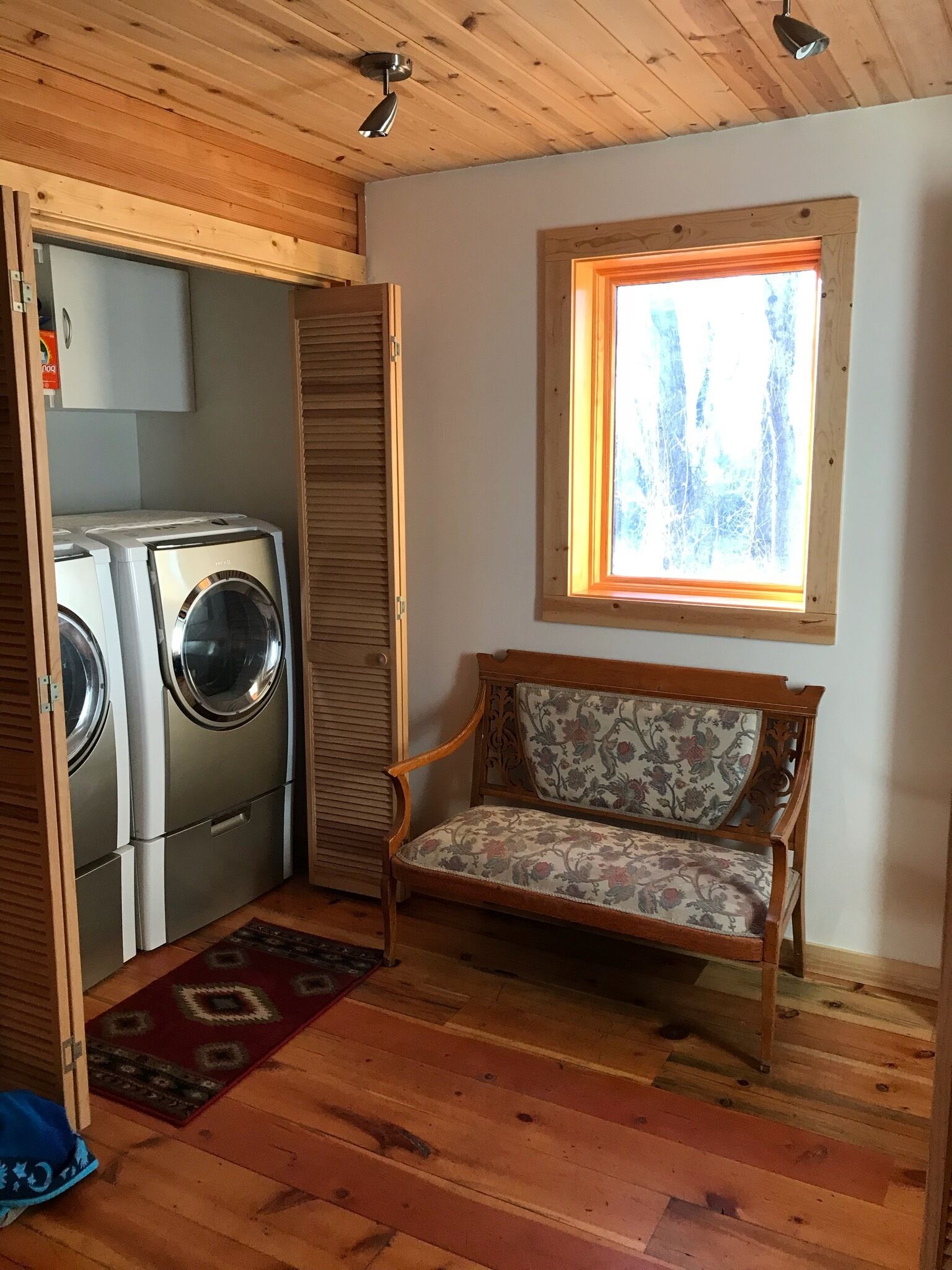 A room with a wooden ceiling, a window with a view of trees, a washing machine, a bench, and a patterned rug.