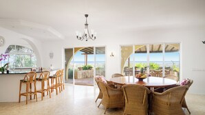 A spacious dining area with wicker chairs and a round table, large windows offering a view of the outdoors, and a kitchen area with bar stools. - Charming home with panoramic sea views, close to Aquashow, Airport & beaches (Almancil)