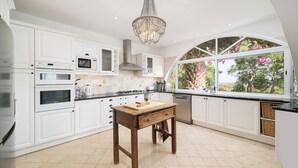 A kitchen with white cabinetry, a wooden table, and a view of trees through a large window. - Charming home with panoramic sea views, close to Aquashow, Airport & beaches (Almancil)