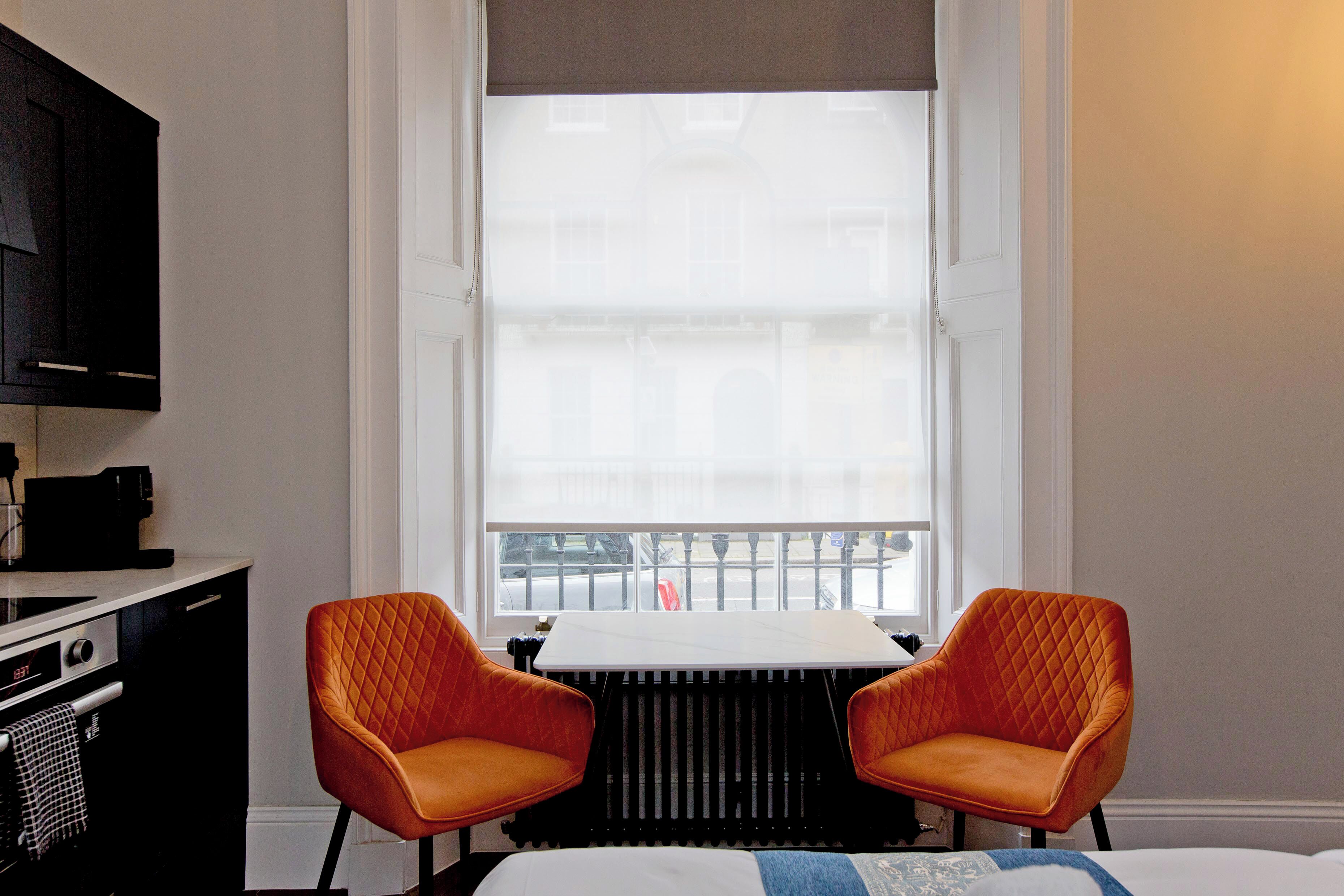 Hotelkamer met een bed, een tafel, twee oranje stoelen, een radiator en een raam met uitzicht op een gebouw.