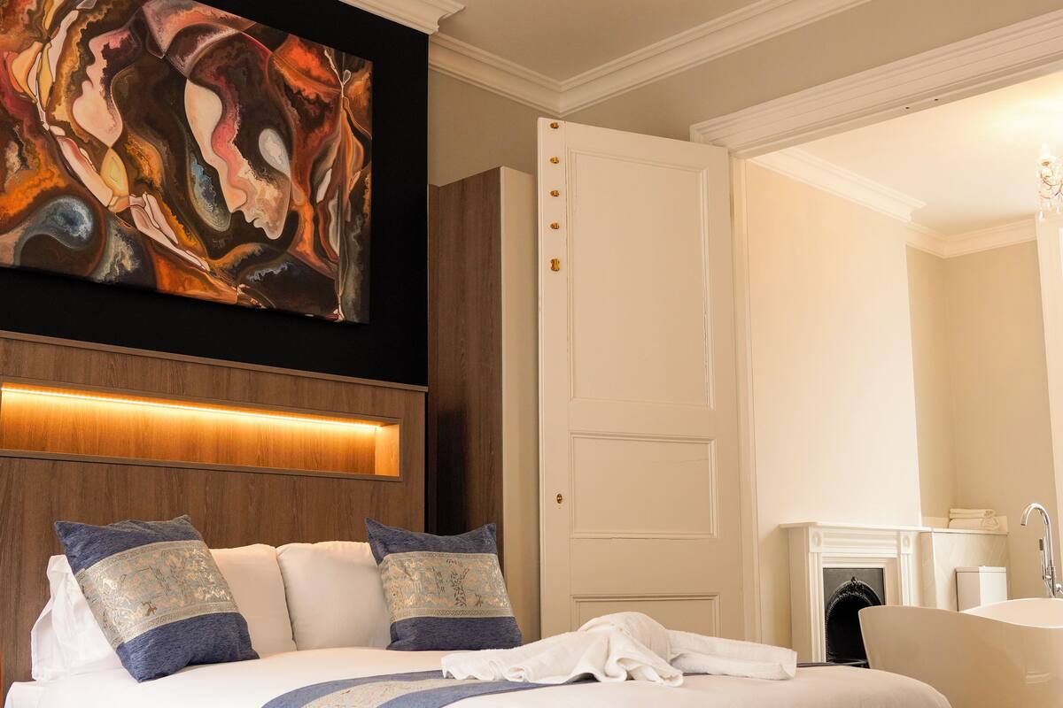 A hotel room with a large bed, a painting above the bed, a wooden panel wall, a white door, and a bathroom with a bathtub and sink.