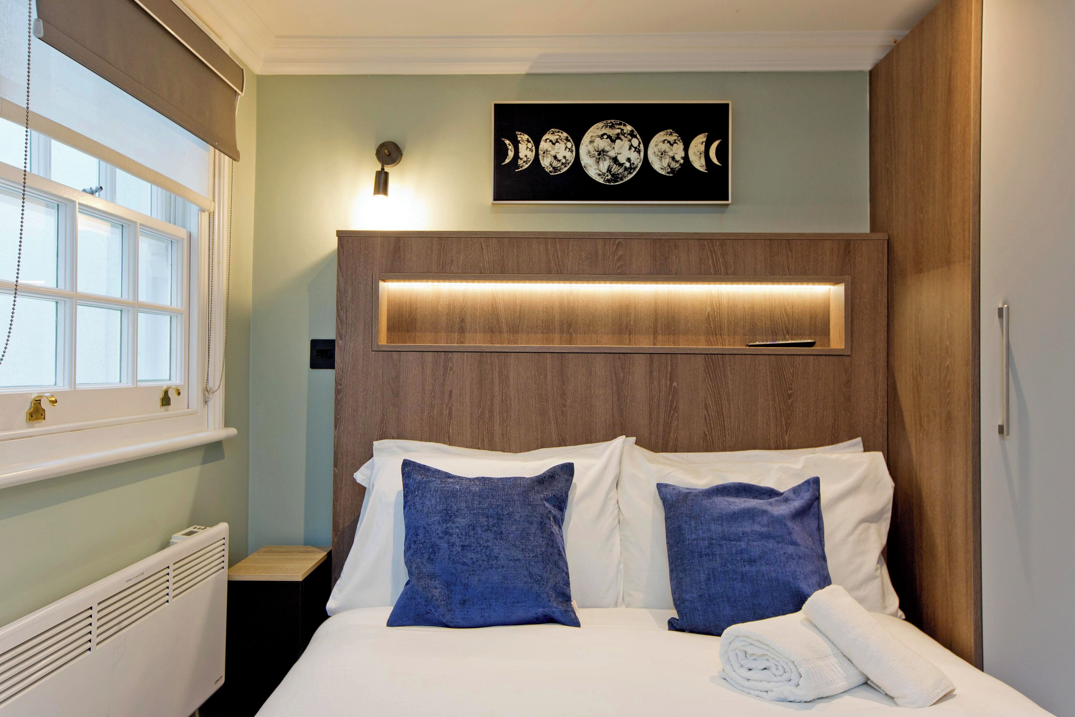 A bedroom with a wooden headboard, a wall-mounted artwork of moon phases, a bedside table, and a rolled towel on the bed.
