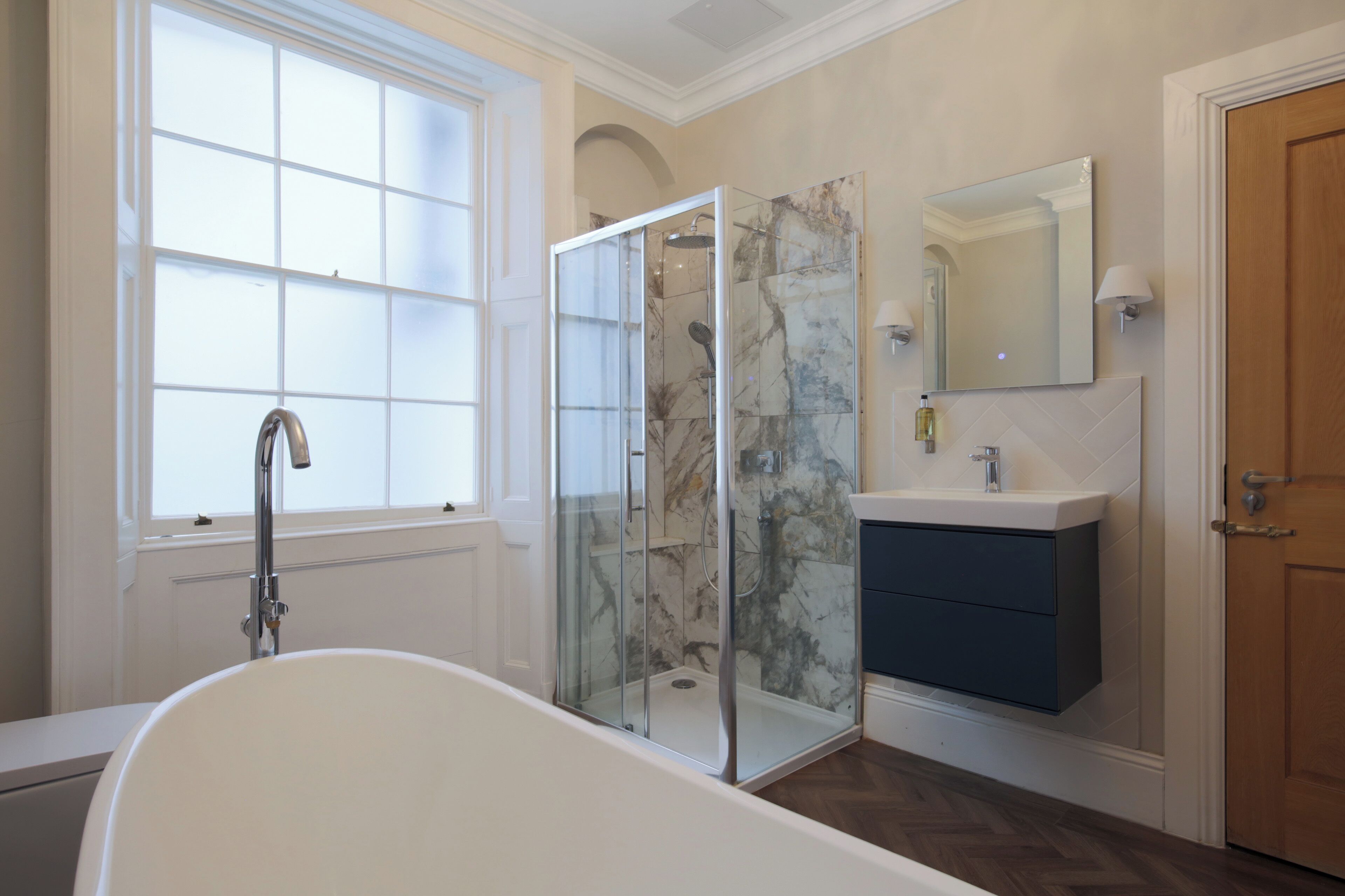 A bathroom with a glass-enclosed shower, a freestanding bathtub, a vanity with a mirror, and a window providing natural light.