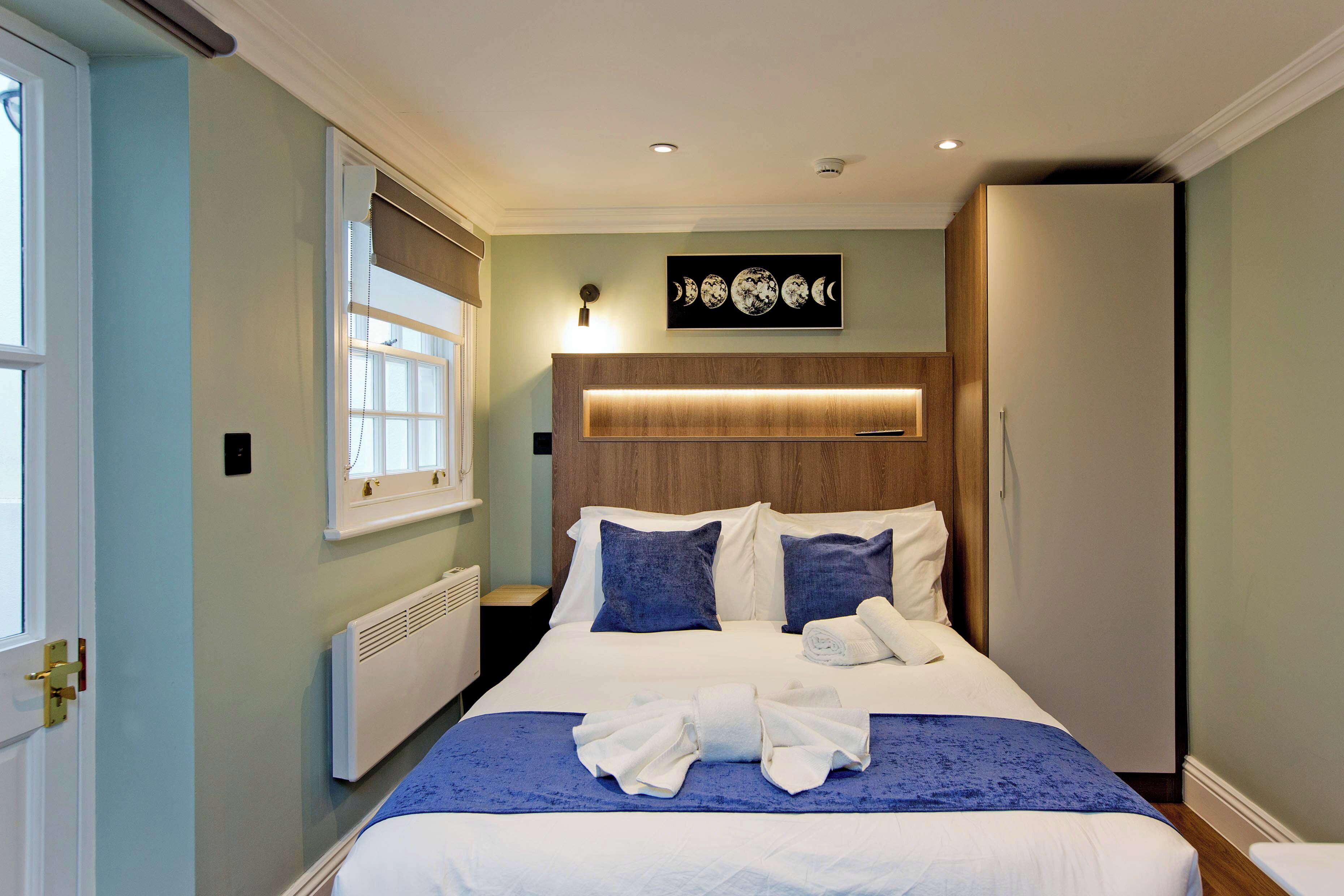 A neatly made bed with a blue bedspread and white linens, a wooden headboard, and a wall-mounted artwork of moon phases.