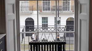 Vista a través de una ventana con baranda metálica, un edificio de ladrillo con toldos rojos y una puerta con marco blanco.