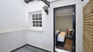 Un pasillo estrecho con una pared blanca, una ventana y una puerta que da a una habitación con una cama, una silla y una mesa.