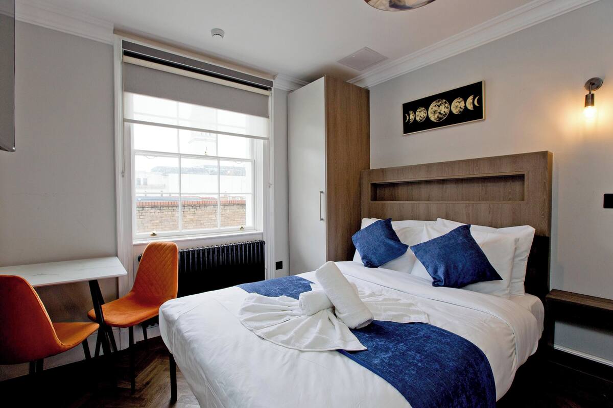 A hotel room with a bed, a desk, a chair, a window, and a wall-mounted artwork.