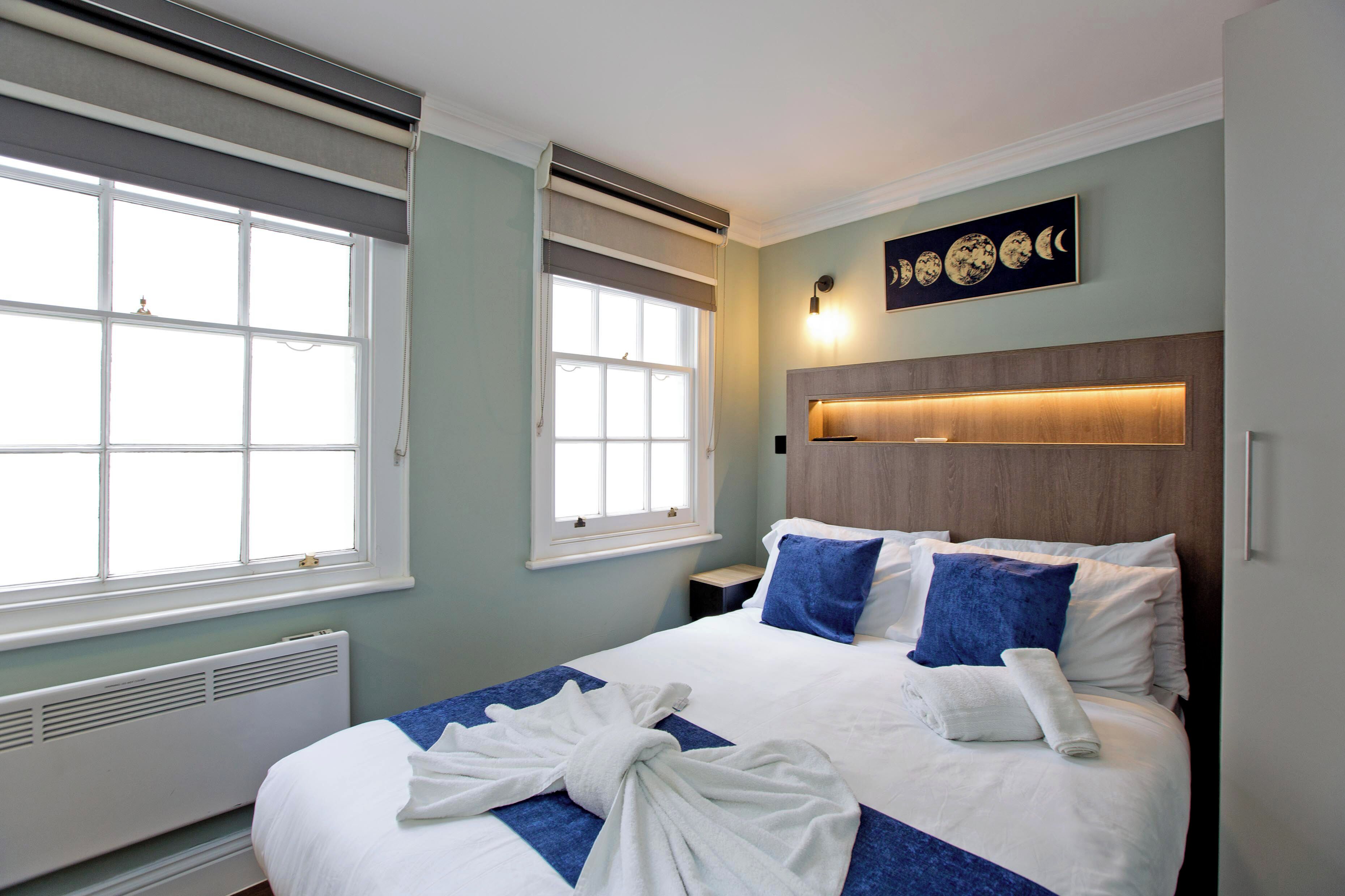 Quarto de hotel com uma cama grande, duas janelas, uma luminária fixada na parede e uma obra de arte com as fases da lua.