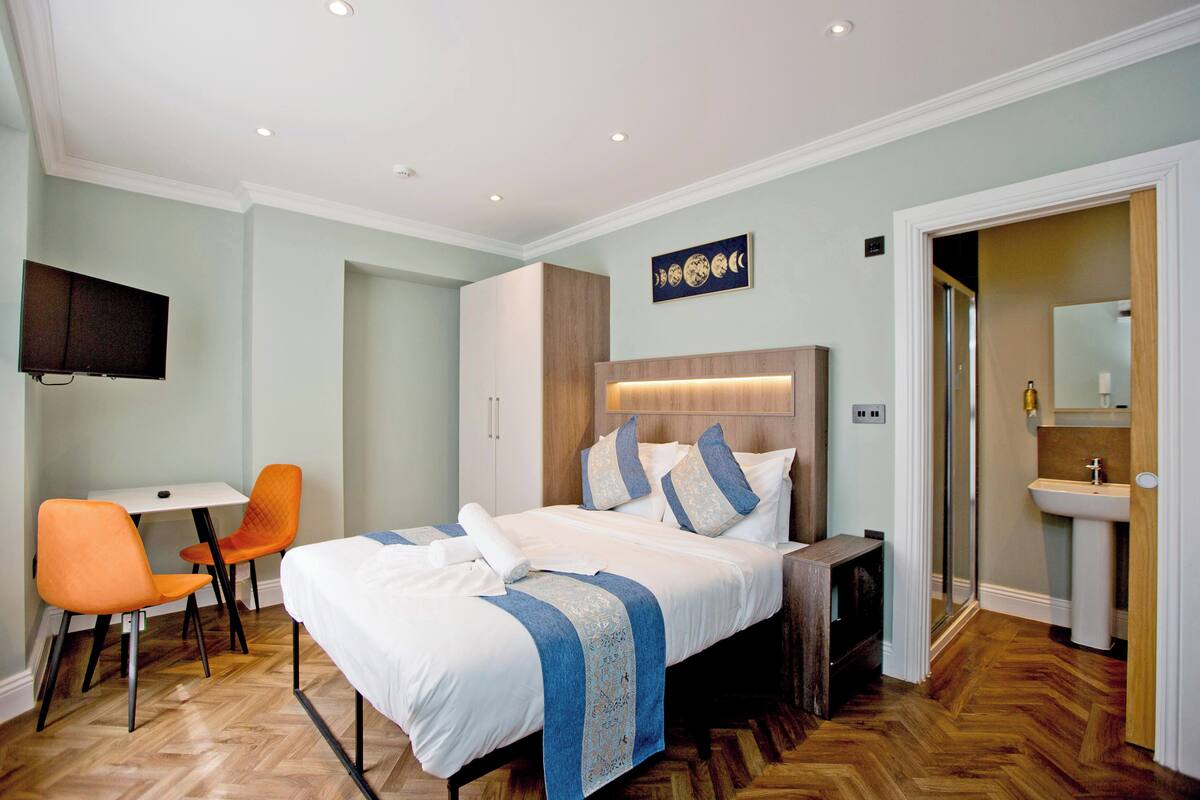 A hotel room with a bed, a desk, an orange chair, a TV, and a bathroom with a sink.