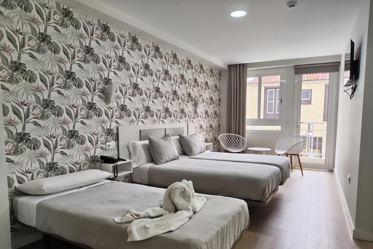 A hotel room with two beds, a small table, a chair, and a balcony with a view.