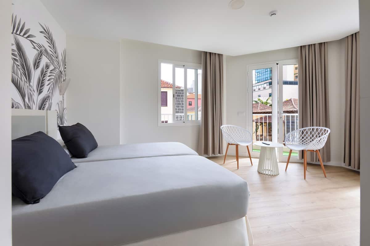 A modern hotel room with a bed, two chairs, a small table, and a view of the city through large windows.