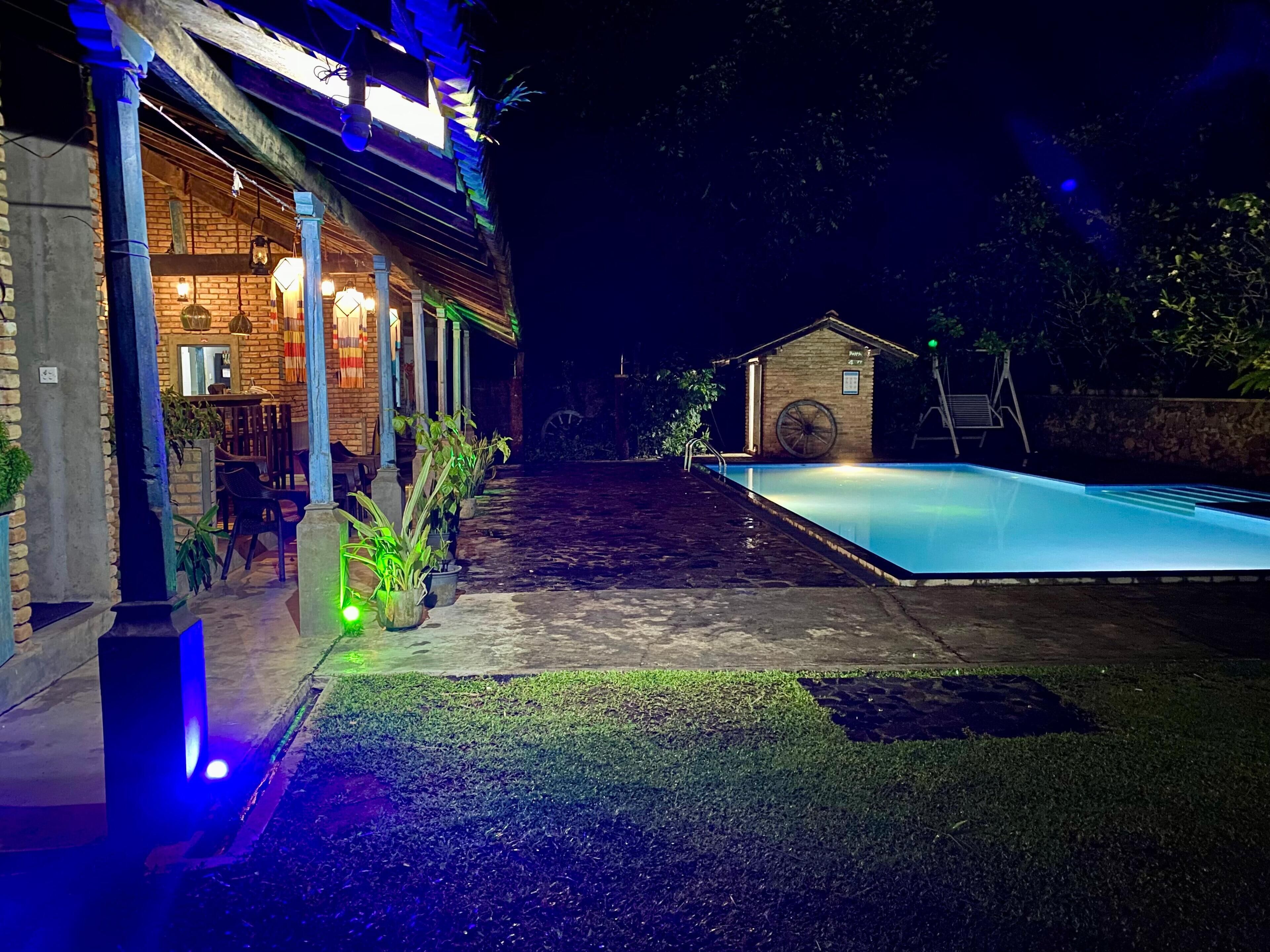 Vista nocturna d'una zona de piscina amb edifici de maó, mobiliari exterior i un petit cobert.