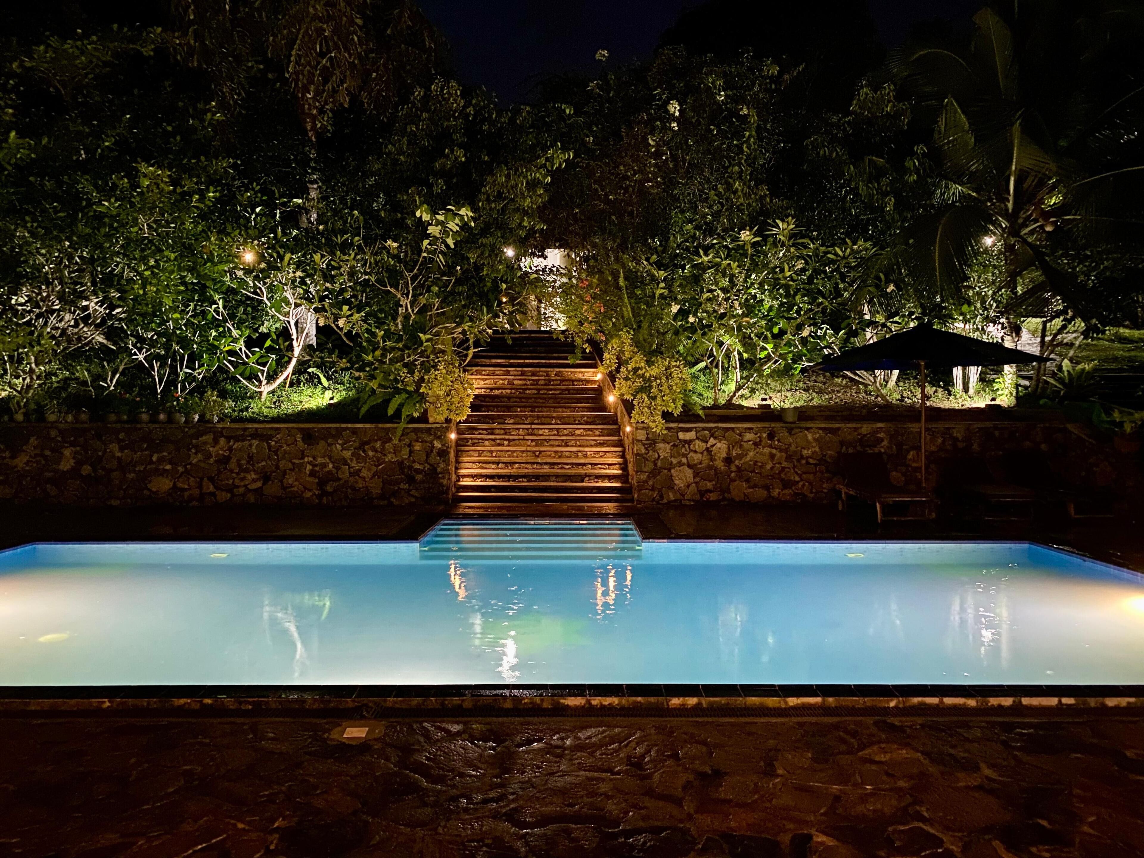 A nighttime swimming pool with a wooden staircase leading up to a stone wall, surrounded by lush greenery.