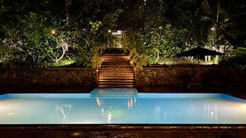 A nighttime swimming pool with a wooden staircase leading up to a stone wall, surrounded by lush greenery.