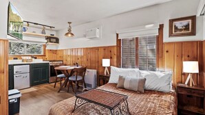 A compact room with a bed, bedside table, lamp, and a small dining table with chairs. There is a kitchen area with a refrigerator, stove, and cabinets. A window provides natural light and a view of greenery outside. - Timber Lodge Cabin #11 (Colorado Springs)