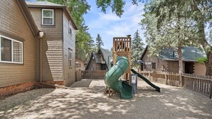 Een speeltuin met een glijbaan en klimconstructie, omringd door houten huizen en bomen.