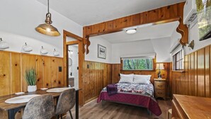 A cozy cabin-style room with a wooden interior, a bed with a quilt, a dining table with chairs, and a window overlooking a forest. - Timber Lodge Cabin #5 (Colorado Springs)