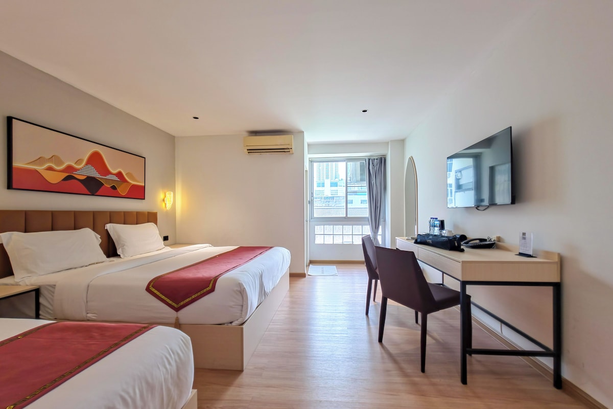 Een hotelkamer met twee bedden, een bureau, een stoel, een televisie en een raam met uitzicht op gebouwen.