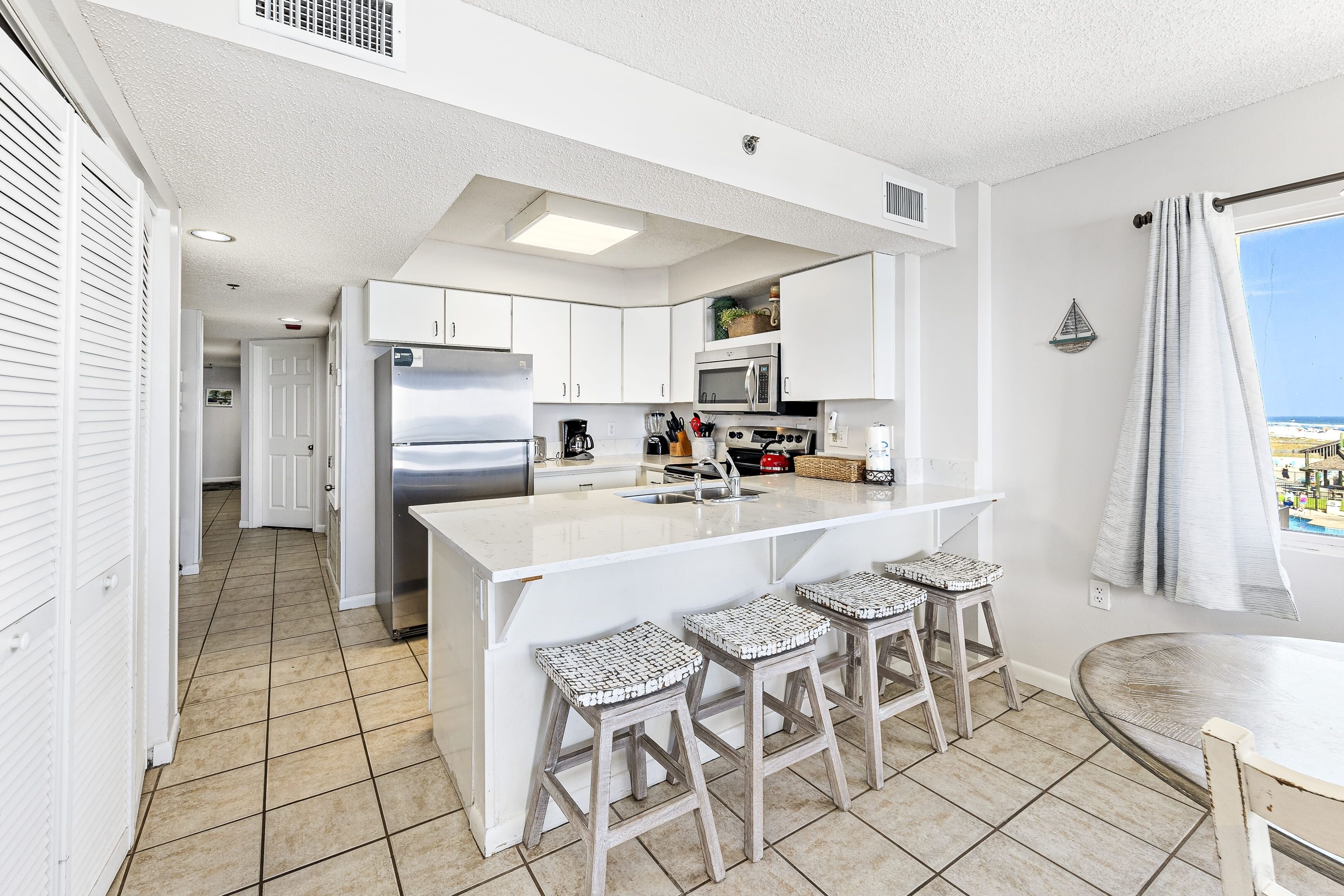 A modern kitchen with white cabinetry, a large island with stools, a stainless steel refrigerator, and a view of the ocean through a window.