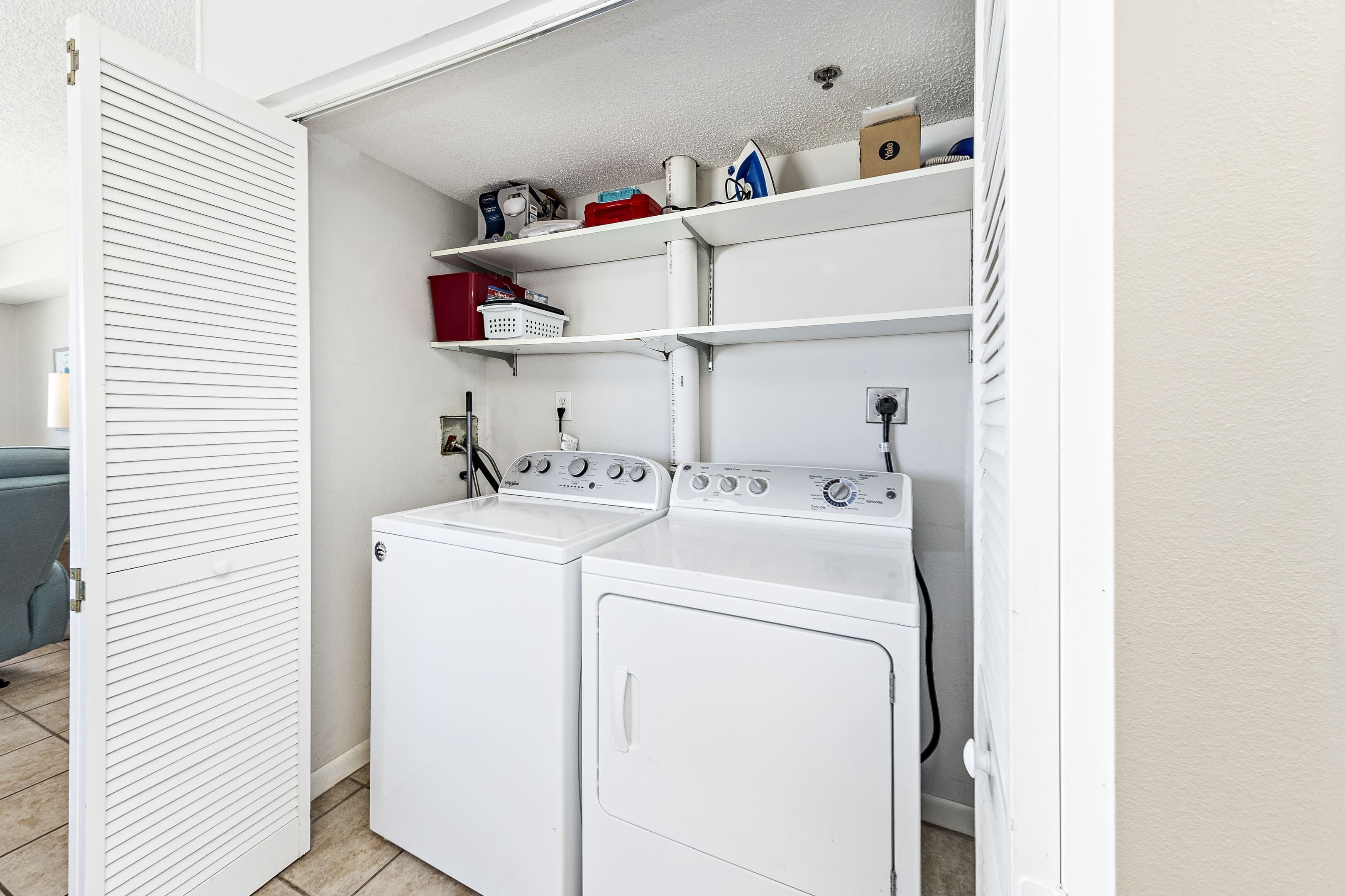 A laundry room with two white front-loading washing machines, a shelf with various items, and a door with horizontal slats.