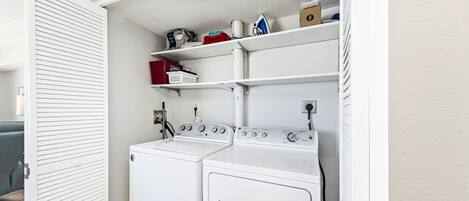 A laundry room with two white front-loading washing machines, a shelf with various items, and a door with horizontal slats.