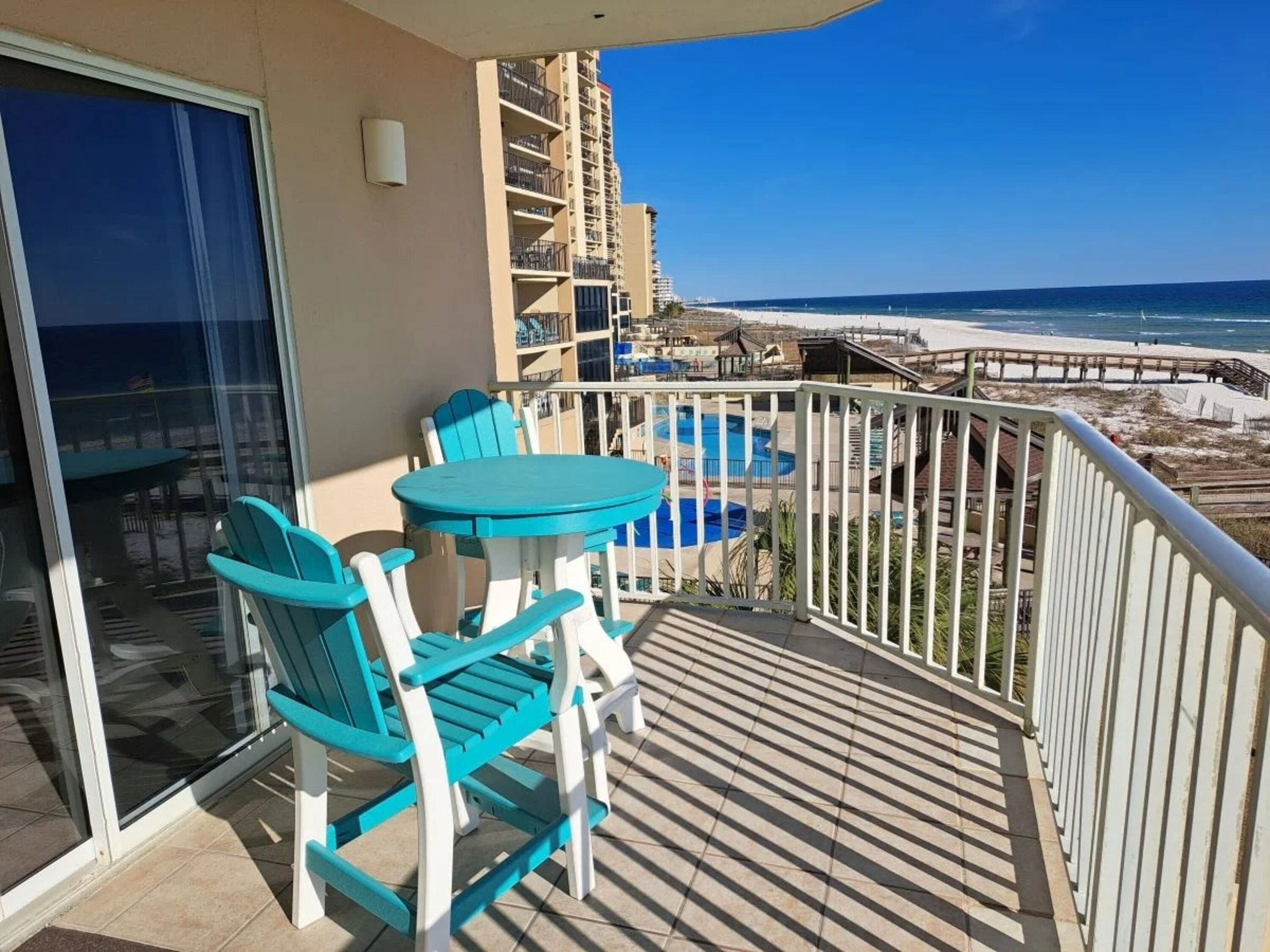 A balcony with a view of the beach and ocean, a pool, and a dining area with chairs and a table.