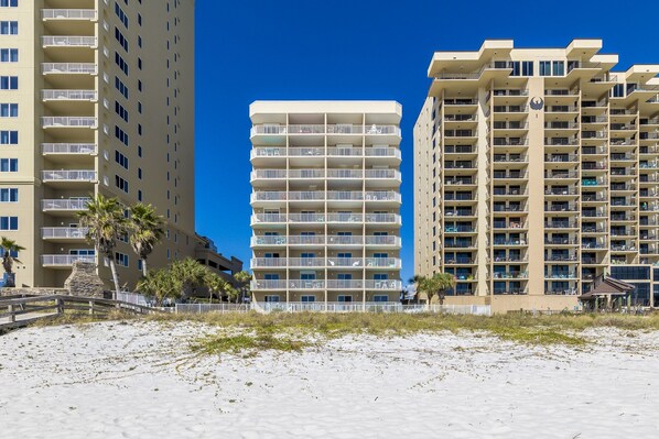 A beachfront hotel with multiple balconies, a sandy beach in the foreground, and palm trees. - Four Winds 301 (Orange Beach)