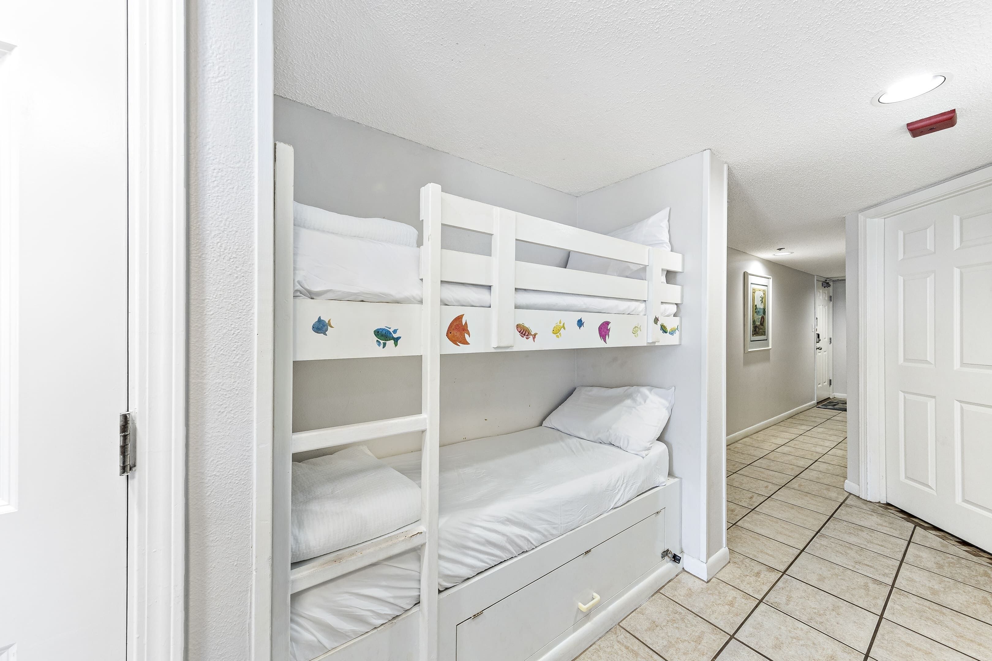 Una cama litera con una plaza inferior y una superior, ambas con ropa de cama blanca. Hay un cajón pequeño debajo de la cama inferior. La habitación tiene paredes blancas y suelo de baldosas.