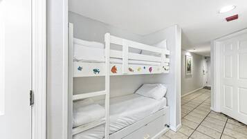 Una cama litera con una plaza inferior y una superior, ambas con ropa de cama blanca. Hay un cajón pequeño debajo de la cama inferior. La habitación tiene paredes blancas y suelo de baldosas.