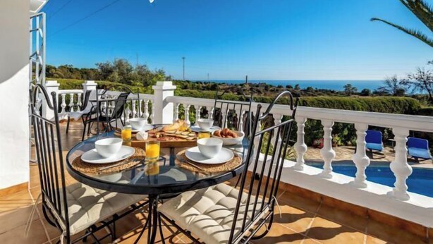 Una mesa redonda con platos y tazones blancos, un vaso de zumo de naranja y una cesta de pan en un balcón con barandilla blanca y vistas al mar.
