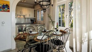 Un comedor con una mesa de vidrio, sillas de metal y una araña. Se aprecia una cocina al fondo con gabinetes blancos y una estufa.