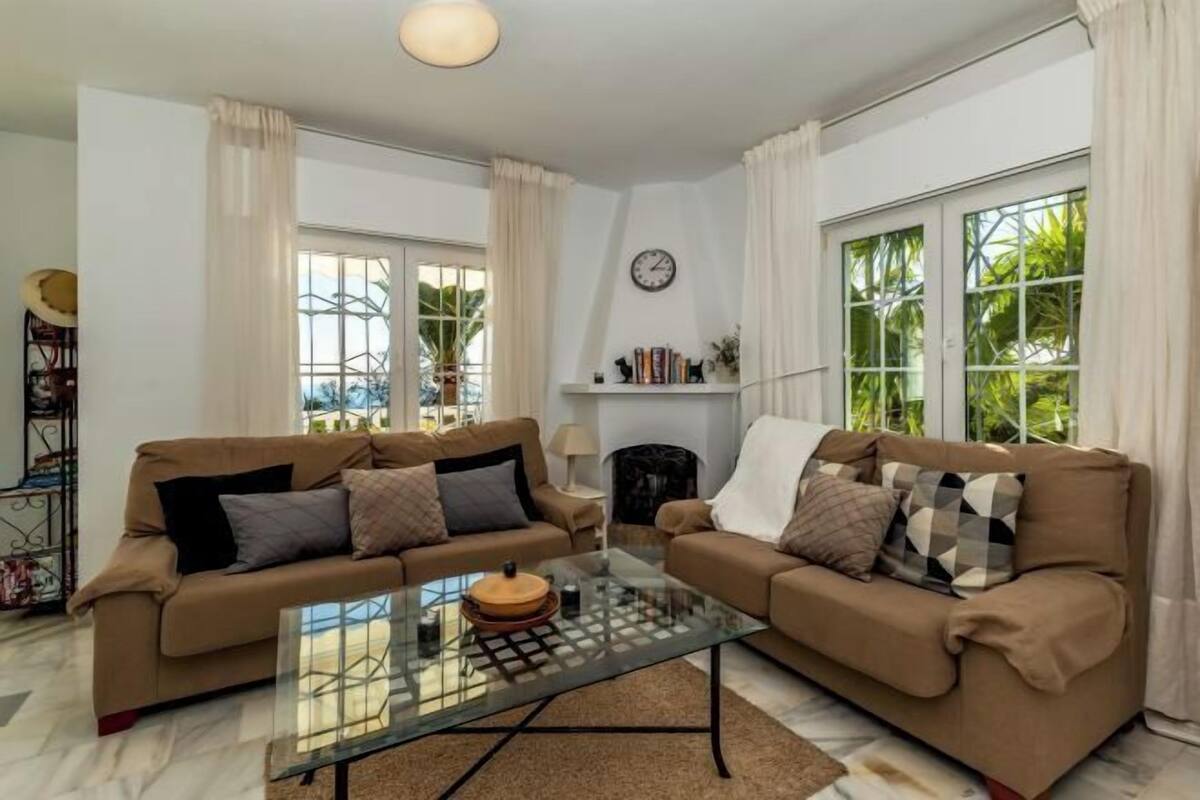 A living room with two sofas, a glass coffee table, a fireplace, and large windows with curtains.