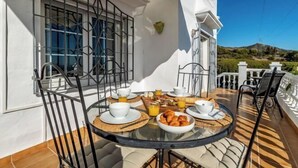 Una mesa redonda con alimentos para el desayuno, como cruasanes, zumo de naranja y tazas de café, en un balcón con pared blanca y barandilla de metal.