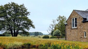 Un edificio de piedra con techo inclinado y una pequeña ventana, rodeado por un campo cubierto de hierba y un estanque con flores amarillas.