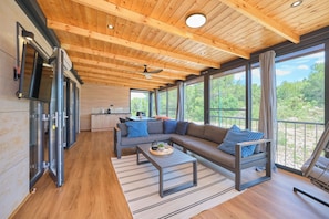 A modern living room with a wooden ceiling, large windows, a sofa, a coffee table, and a kitchen area.