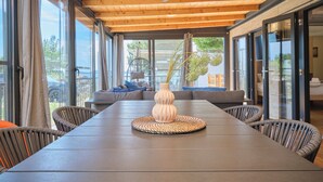 A modern dining area with a long table, wicker chairs, a ceiling fan, and large windows offering a view of the outdoors. - MH Holiday Dream - Shining Sea - Oaza Mira campsite (Drage)
