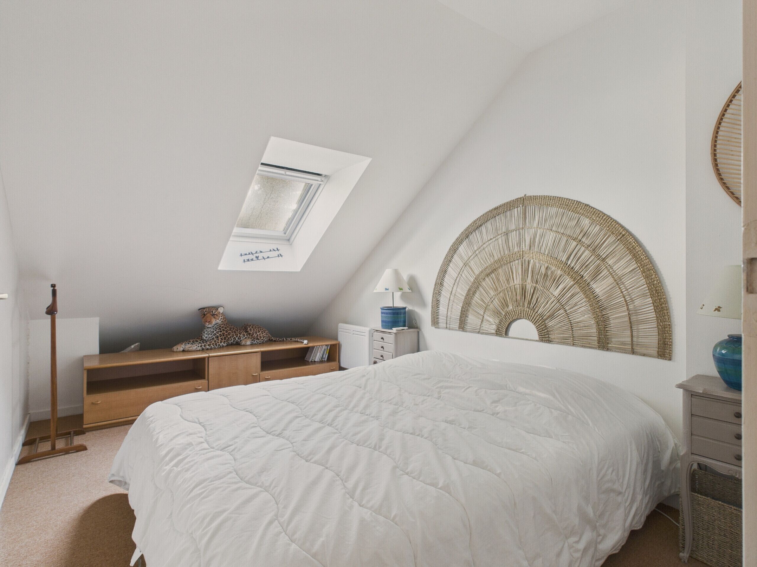 A bedroom with a sloped ceiling, a bed with a white duvet, a wooden bench, a small table with a lamp, and a decorative wall piece.
