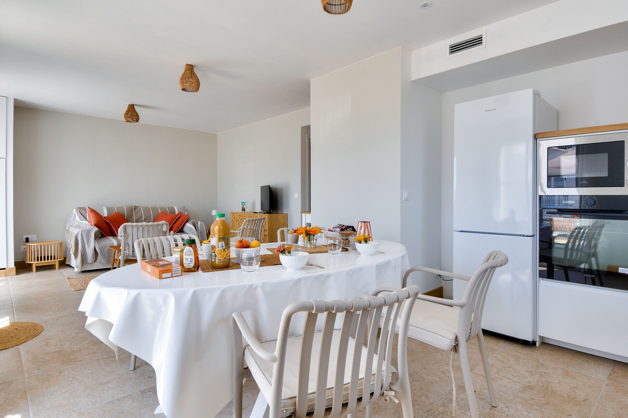 Un espace repas moderne avec une table ronde dressée pour quatre personnes, entourée de chaises blanches. Un réfrigérateur et un four blancs sont visibles à l’arrière-plan.