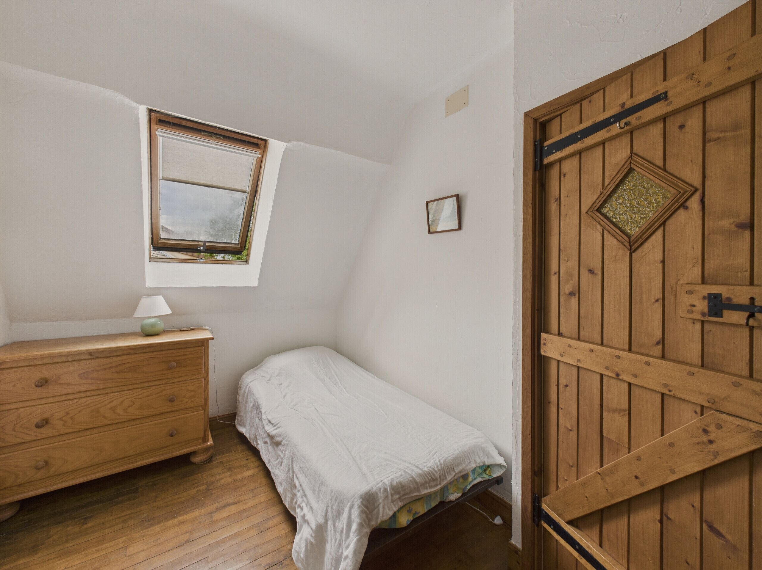 Una habitación pequeña con cama individual, un aparador de madera, una ventana con vistas y una puerta de madera con un ventanal en forma de rombo.