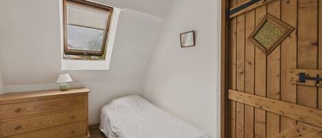 Una habitación pequeña con cama individual, un aparador de madera, una ventana con vistas y una puerta de madera con un ventanal en forma de rombo.