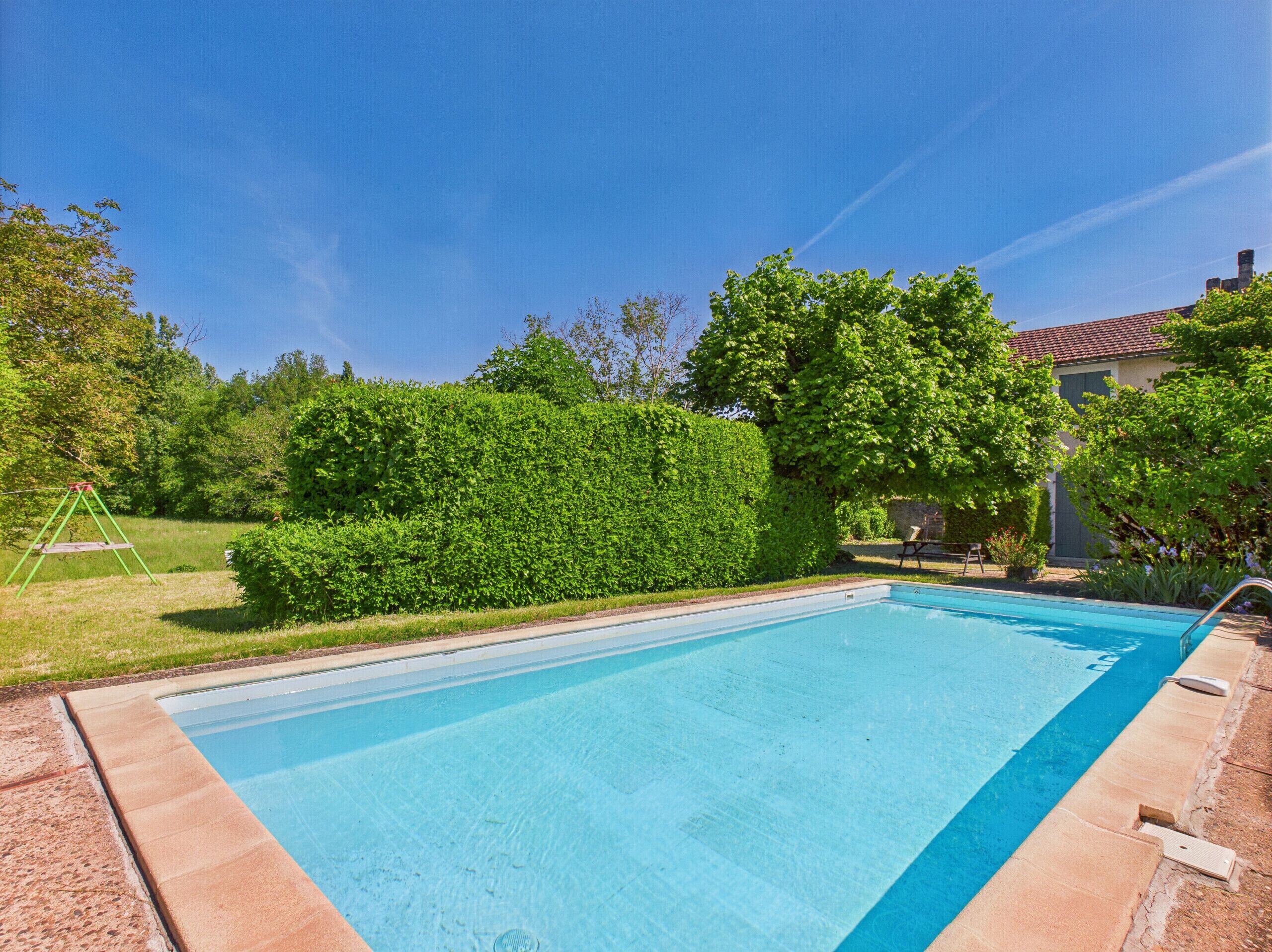 A rectangular swimming pool with a clear blue water surface, surrounded by a concrete deck and a low hedge.